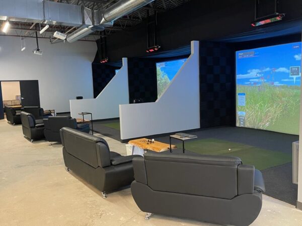 The image shows the interior of an indoor golf facility featuring two golf simulators, each with a large screen displaying a virtual golf course. The simulators are separated by white walls, and in front of each, there are black leather couches arranged in pairs for seating. Small wooden tables are positioned in front of the couches, and overhead, various lighting and ventilation systems are visible. The setting appears modern and comfortable, designed for both playing and relaxation